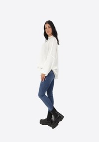 Maglione bianco oversize con fessure laterali, indossato con jeans skinny blu e stivaletti neri con tacco largo. Tessuto morbido e vestibilità rilassata.