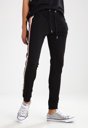 Persoon draagt zwarte joggingbroek met regenboogkleurige strepen aan de zijkant en zilveren lage sneakers, staand tegen een witte achtergrond.