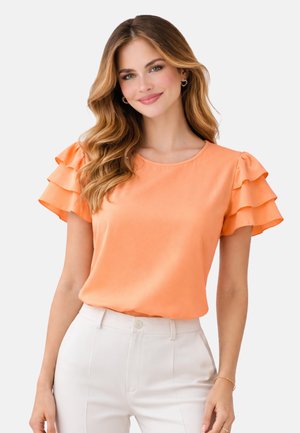 Kobieta o falujących włosach, ubrana w pomarańczową bluzkę z warstwowymi falbanami na rękawach oraz białe, wysokie spodnie, uśmiecha się na jednolitym tle.