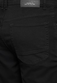 Schwarze Jeans aus strukturiertem Stoff, mit einer Gesäßtasche mit abgerundeten Kanten und einem Markenlabel darüber. Dezente Steppnähte sind sichtbar.