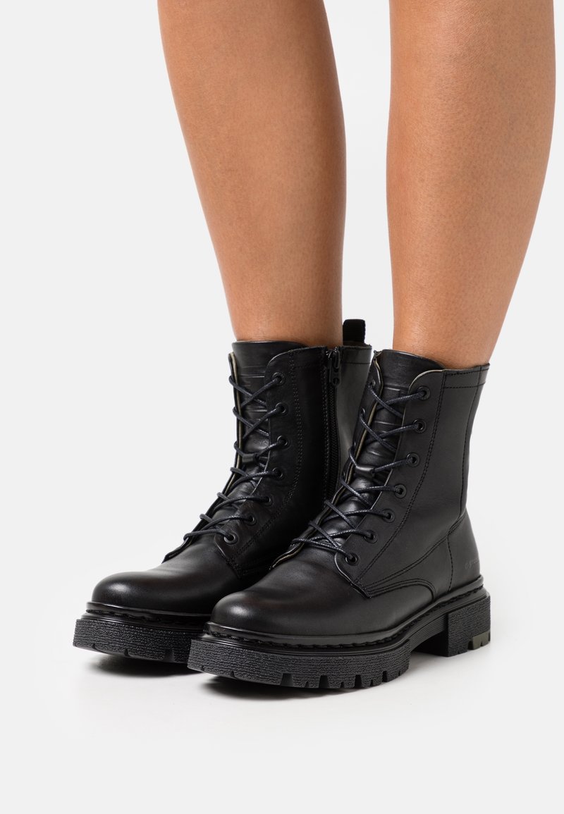 Personne portant des bottes de combat noires à lacets avec des semelles épaisses crantées, debout sur un fond blanc.
