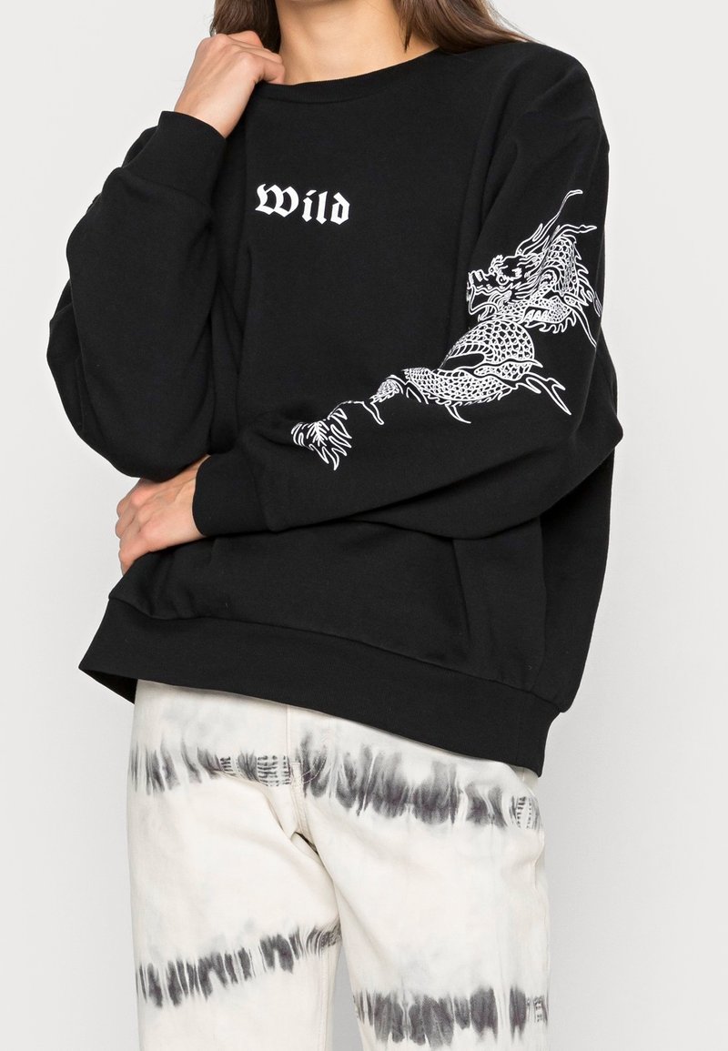 Sweat-shirt noir avec le mot "Wild" sur la poitrine et un motif de dragon blanc enroulé autour de la manche droite, associé à un pantalon tie-dye blanc et gris.