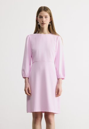 Mladá žena v svetloružových, dlhých rukávoch až po kolená s priliehavým pásom a náušnicami s perlami, stojaca pred jednoduchým pozadím.