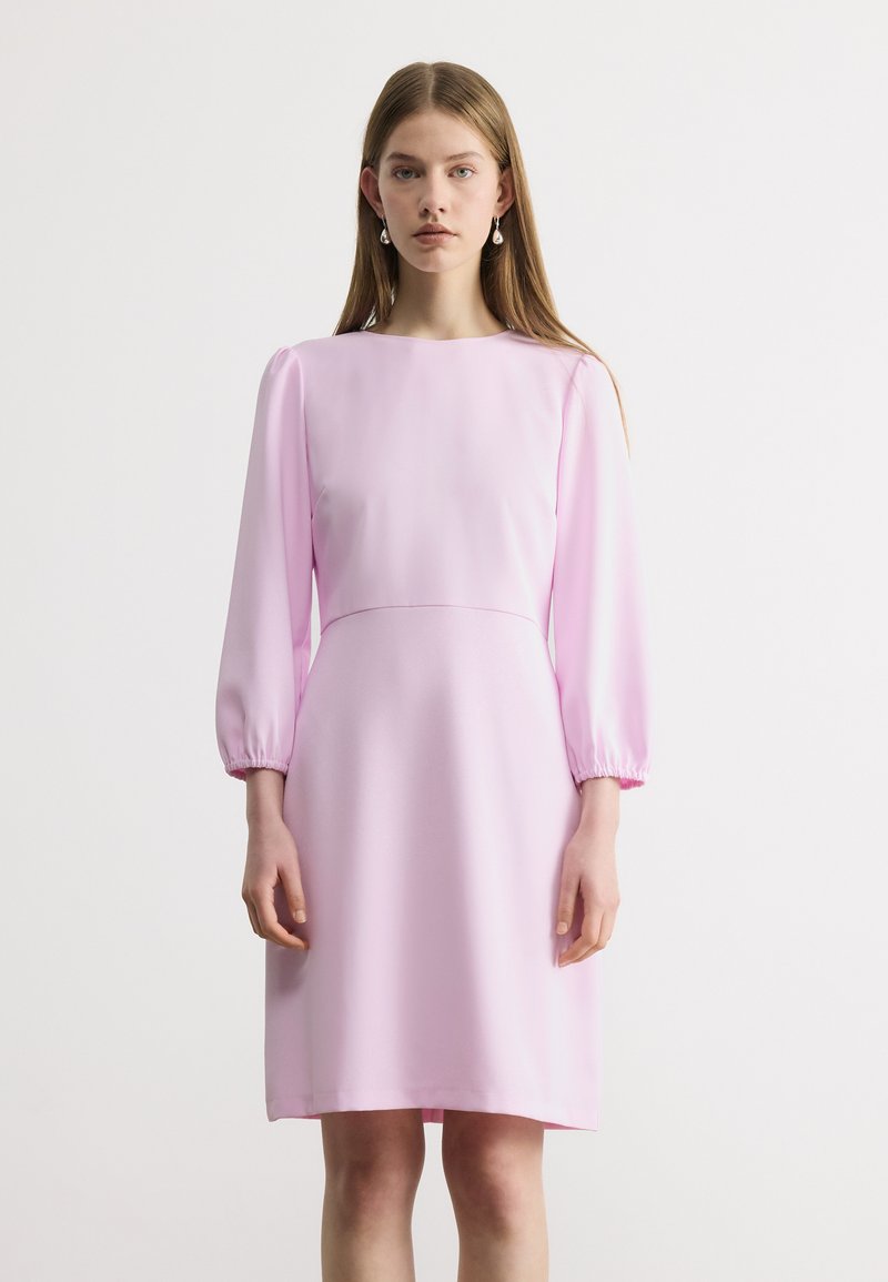 Jeune femme portant une robe rose pâle à manches longues, longueur genou, taille cintrée et boucles d'oreilles pendantes en perles, debout devant un fond uni.