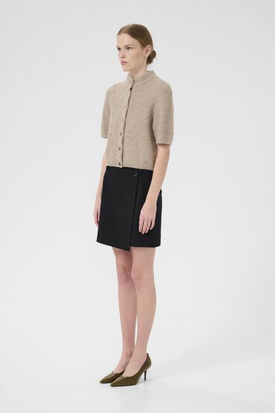 Cardigan en tricot beige à manches courtes avec encolure ronde et boutons, associé à une jupe noire en A avec fermeture à bouton sur le côté. Escarpins oliviers.