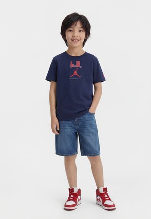Niño con cabello negro, vestido con camiseta gráfica azul marino de L.A., pantalones cortos de mezclilla y zapatillas rojas y blancas, de pie con una mano en el bolsillo.