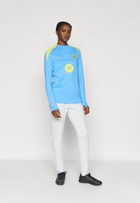 Γαλάζια αθλητική μπλούζα με μακριά μανίκια και κίτρινα στοιχεία, στρογγυλό λογότυπο και φερμουάρ στον λαιμό· σε συνδυασμό με λευκό παντελόνι και μαύρα αθλητικά παπούτσια.