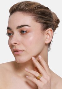 Anello in metallo tono oro con bande testurizzate, indossato sull'indice, abbinato a un tono di pelle neutro. Il modello ha un trucco leggero e i capelli raccolti in uno chignon.