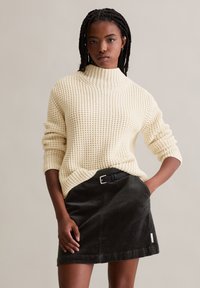 Cremefarbener, gerippter Rollkragenpulli, kombiniert mit einem schwarzen Cordrock, der einen sichtbaren Gürtel und aufgerollte Ärmel für zusätzliche Details aufweist.