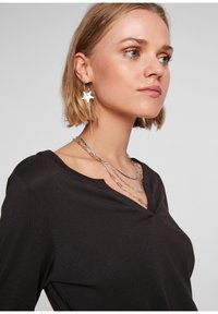 Blonde Frau, die ein schwarzes V-Ausschnitt-Oberteil, geschichtete Silberketten und silberne, sternförmige Creolen trägt und nach rechts schaut.