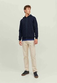 Homemem de pé, vestindo um cardigan navy com fecho, calças bege e ténis escuros, olhando para o lado contra um fundo claro e simples.