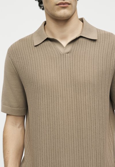 Άνδρας που φοράει μπεζ κοντομάνικο πλεκτό πόλο με υφασμένο κάθετο μοτίβο και γιακά με V-κοπή.