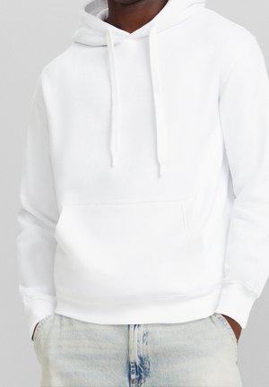 Homme portant un sweat à capuche blanc uni avec poche avant et cordons, associé à un jean bleu clair, les mains dans les poches.