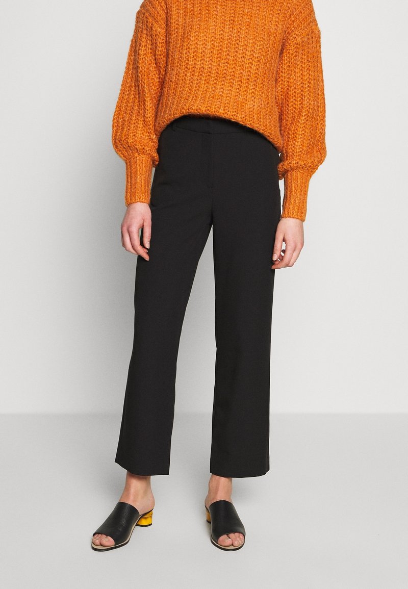 Oranje gebreide trui met een losse pasvorm en ribbelstructuur, gecombineerd met zwarte wijde broek. Zwarte open-teenschoenen met gele accenten.