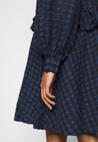 Sininen ja musta ruudullinen mekko, jossa on volangiyksityiskohta, teksturoitu kangas, puffihihat ja napitettu hihansuu, esittäen rentoa istuvuutta.