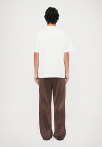 T-shirt blanc à manches courtes avec col rond, associé à un pantalon marron ample. Le vêtement semble fait de tissu doux et léger.