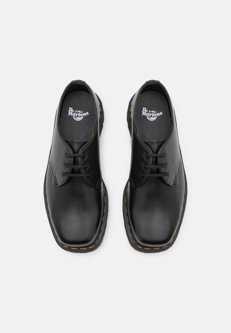 Zapatos de cuero negros con punteras redondeadas, dos ojetes para los cordones, costuras amarillas a lo largo de la suela, plantillas texturizadas, diseño clásico.