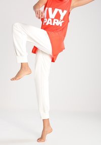 Rød ærmeløs top med "IVY PARK" i store hvide bogstaver, stylet med elfenbenfarvede joggerbukser. Outfittet har en løs pasform og blødt stof.