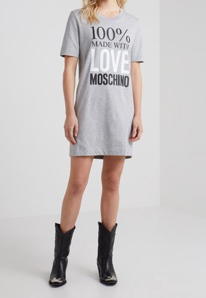 Kvinde iført en grå kjole med korte ærmer med teksten "100% Made with Love Moschino", kombineret med sorte støvler til midt på læggen.