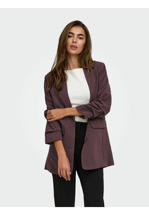 Donna che indossa un top bianco, blazer viola scuro con le maniche arrotolate e pantaloni neri, in piedi davanti a uno sfondo grigio chiaro.