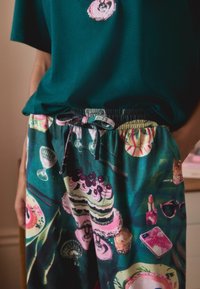 Zielona bawełniana bluzka z okrągłym dekoltem w połączeniu z kolorowymi spodniami w wzory deserów i dodatków; elastyczny pas ze sznureczkiem do regulacji.