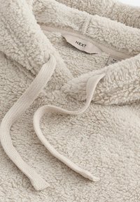 Vaalean beige fleece-huppari, jossa on teksturoitu pinta, varustettu kiristysnauhalla ja brändilogo "NEXT" näkyvissä sisällä.