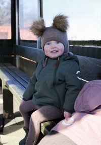 Giacca impermeabile verde con zip, cappello marrone lavorato a maglia con due soffici pon pon, leggings marroni, seduta su una panchina di legno.