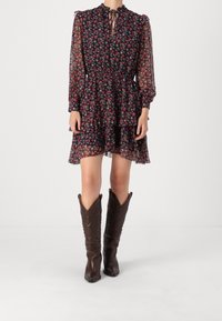 Kvinna i en marinblå blommig långärmad klänning med lagerad kjol och bruna spetsiga cowboystövlar som står på en vit bakgrund.