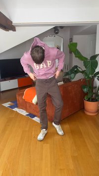 Άτομο με ροζ φούτερ GAP που προσαρμόζει καφέ παντελόνι, στέκεται σε ξύλινο πάτωμα σε καθιστικό, όπου διακρίνονται καναπές, τηλεόραση και μεγάλο γλαστρικό φυτό.