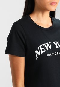 Černé bavlněné tričko s krátkým rukávem, na přední straně s bílým grafickým textem "NEW YORK HILFIGER" ve výrazném, klenutém písmu.