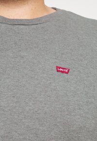 Gros plan sur un t-shirt gris à col rond avec un petit logo rouge Levi's sur le côté gauche de la poitrine, porté par une personne.