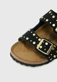 Sort ruskindsandal med guldnitter, to justerbare remme og en formet brun fodseng. Tekstureret gummisål for bedre greb.