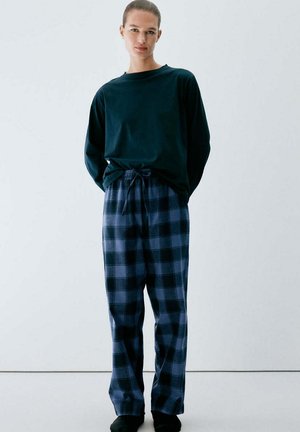 Standing person wearing a dark long-sleeve shirt and blue and black checkered loose pants against a plain light background.