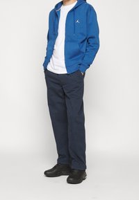 Sudadera azul real con cremallera y logo blanco, camiseta blanca debajo, pantalones oscuros navy y zapatillas deportivas negras. Atuendo casual.