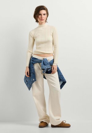 Giovane donna che indossa un dolcevita crema a coste, pantaloni crema a gamba larga, scarpe marroni e una giacca di jeans legata in vita su uno sfondo semplice.