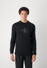 Juodas megztinis su raukšlėta kaklo ir rankovių apdaila. Priekyje yra logotipas su užrašu "Calvin Klein Jeans" pilka ir juoda spalvomis.