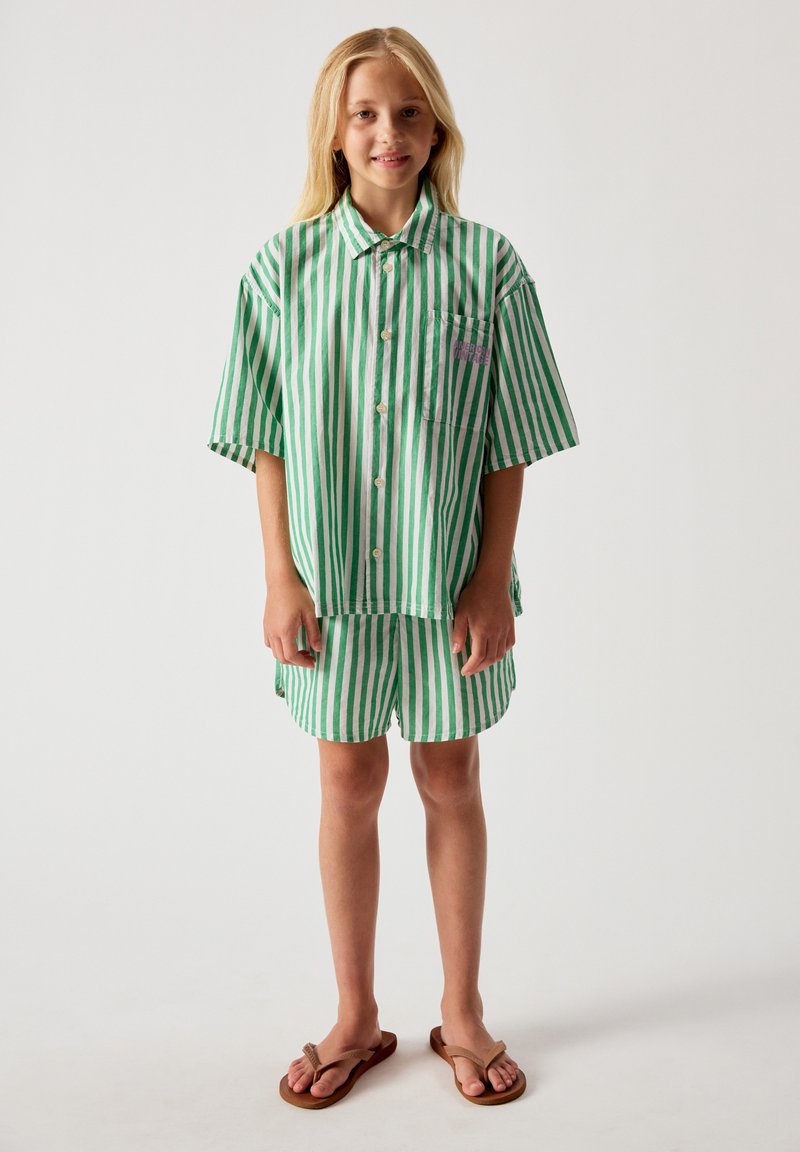 Enfant aux longs cheveux blonds portant une chemise oversize rayée verticale verte et blanche ainsi que des shorts assortis avec des tongs marron.