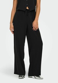 Pantalons larges noirs en tissu doux avec une taille élastique à cordon, associés à des chaussures argentées brillantes. Design minimaliste sans motifs.