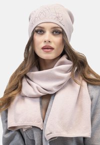 Berretto in maglia rosa chiaro con design ricamato; sciarpa rosa chiaro coordinata con consistenza liscia, indossata sopra un cappotto grigio. Dettaglio in metallo argentato sulla sciarpa.