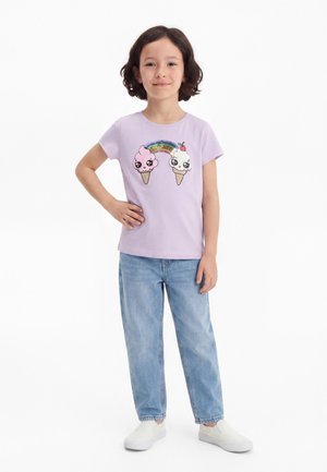 Jente med kort brunt hår står med hånden på hoften, iført lavendelfarget t-skjorte med smilende iskremkjeks og regnbue, lyseblå jeans, hvite sko.