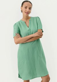 Grünes Kurzarmkleid aus leichtem Material, mit V-Ausschnitt, seitlichen Taschen und einer lockeren Passform mit abgerundetem Saum.