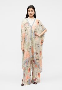 Nő áramló, virágmintás áttetsző köpenyben és hozzá illő, széles szárú nadrágban, fehér garbó és sötét cipő fölött.