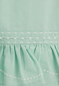 Tissu vert clair avec détails de broderie blanche en point de croix et point courant au-dessus d'une section froncée à volants.