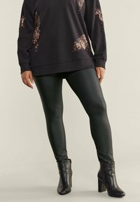 Zwarte sweatshirt met sequins, gecombineerd met glanzende groene leggings en enkellaarsjes. De sweatshirt heeft een relaxed fit en lange mouwen.