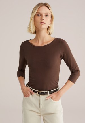 Top marrón de manga larga con escote redondo, hecho de una tela suave y lisa. Combinado con pantalones de color claro y un cinturón negro.