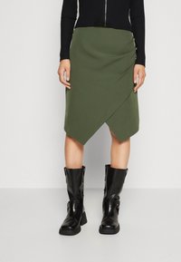 Jupe en olive avec ourlet diagonal et détail de fermeture éclair sur le côté, associée à des bottes noires hautes en matériau brillant.