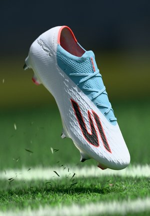 Botte de football blanche avec une surface texturée, des accents bleus et un logo noir. Présente des crampons visibles et un éclaboussement de débris d'herbe.
