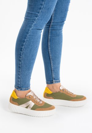 Person wearing blue skinny jeans and green, brown, yellow sneakers with thick white soles, standing on a white background.