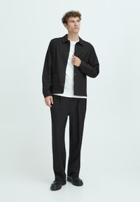 Black button-up jacket with pockets, worn over a white t-shirt; paired with black pleated trousers and black shoes, set against a light background.