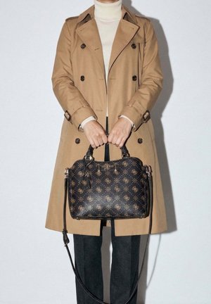 Personne portant un trench beige et un col roulé blanc, tenant un sac à main noir à motif avec deux poignées et une bandoulière.